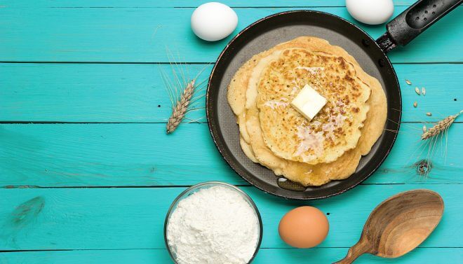 Hot delicious pancakes in frying pan on turquoise wooden table with flour and eggs.