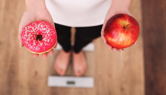 Woman Measuring Body Weight On Weighing Scale Holding Donut and apple.